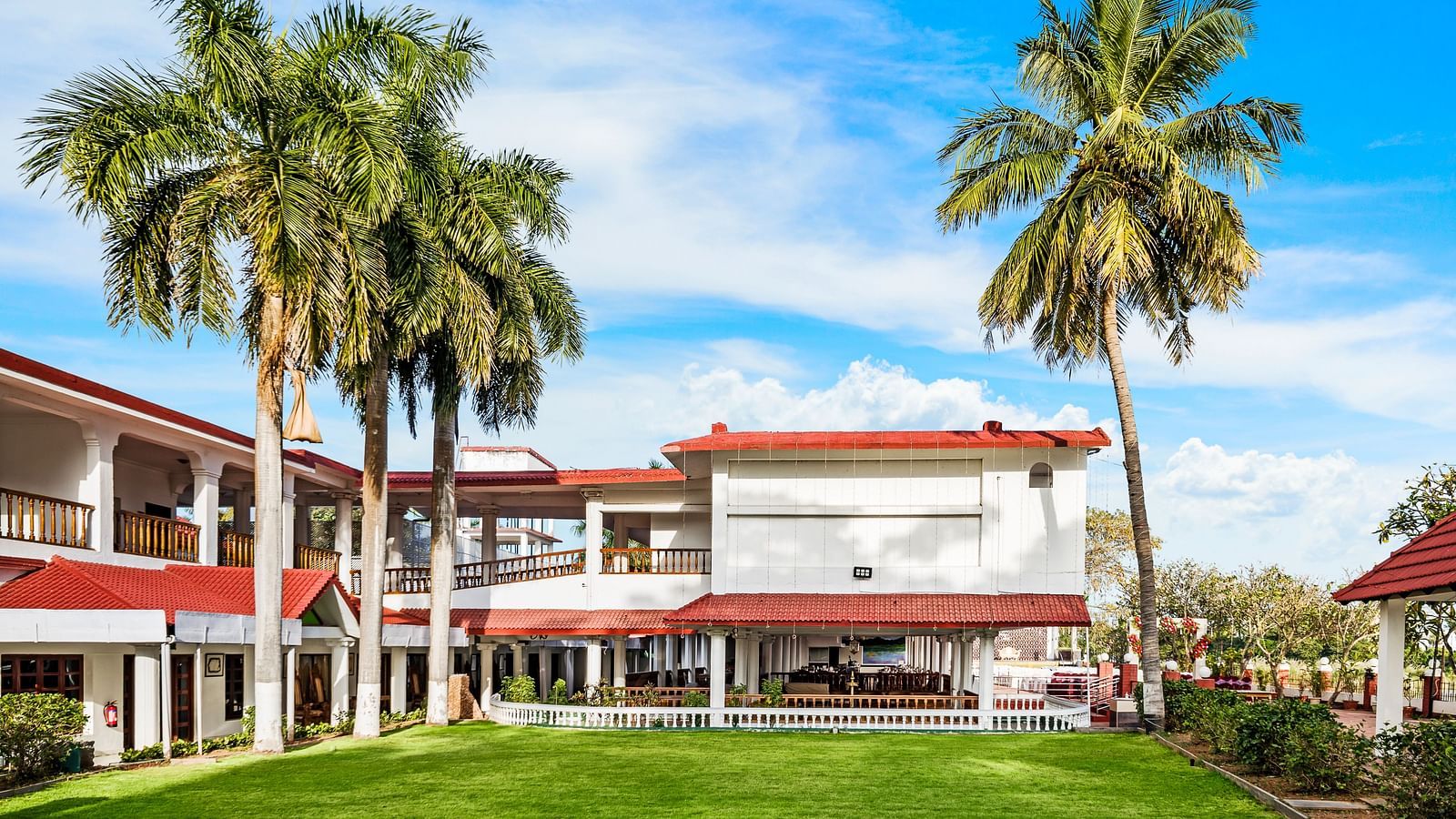 A facade image of the exterior view showcasing a green lawn, tall palm trees with the hotel infrastructure in the background - GReaT Trails River View Resort Thanjavur By GRT Hotels