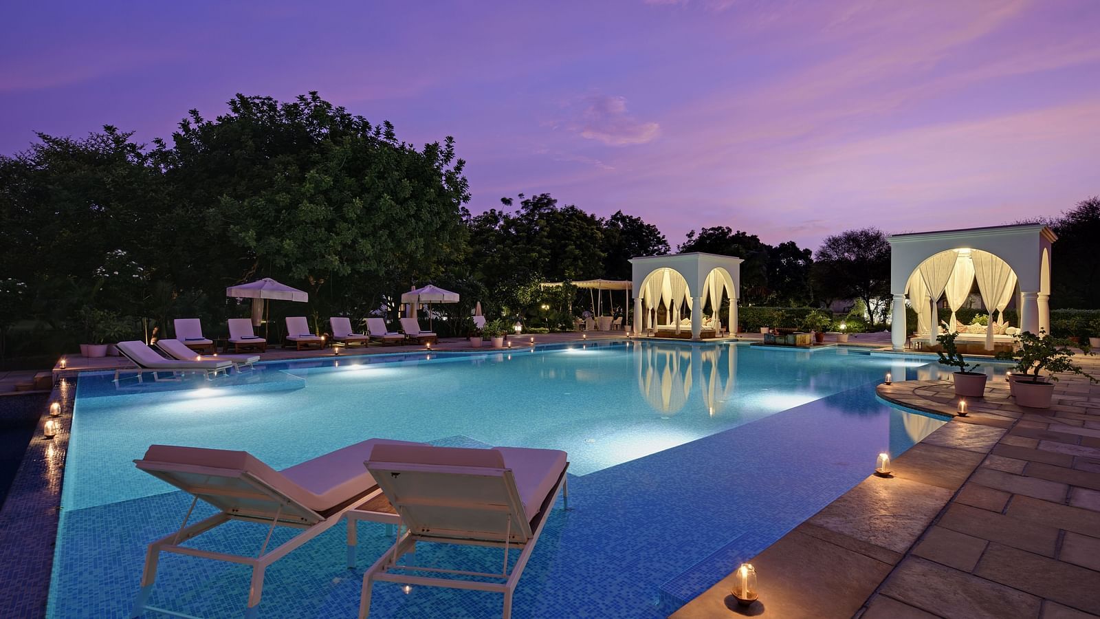 A serene swimming pool at dusk, flanked by lounge chairs and white structures with draped fabric, under a twilight sky- Shahpura Bagh.