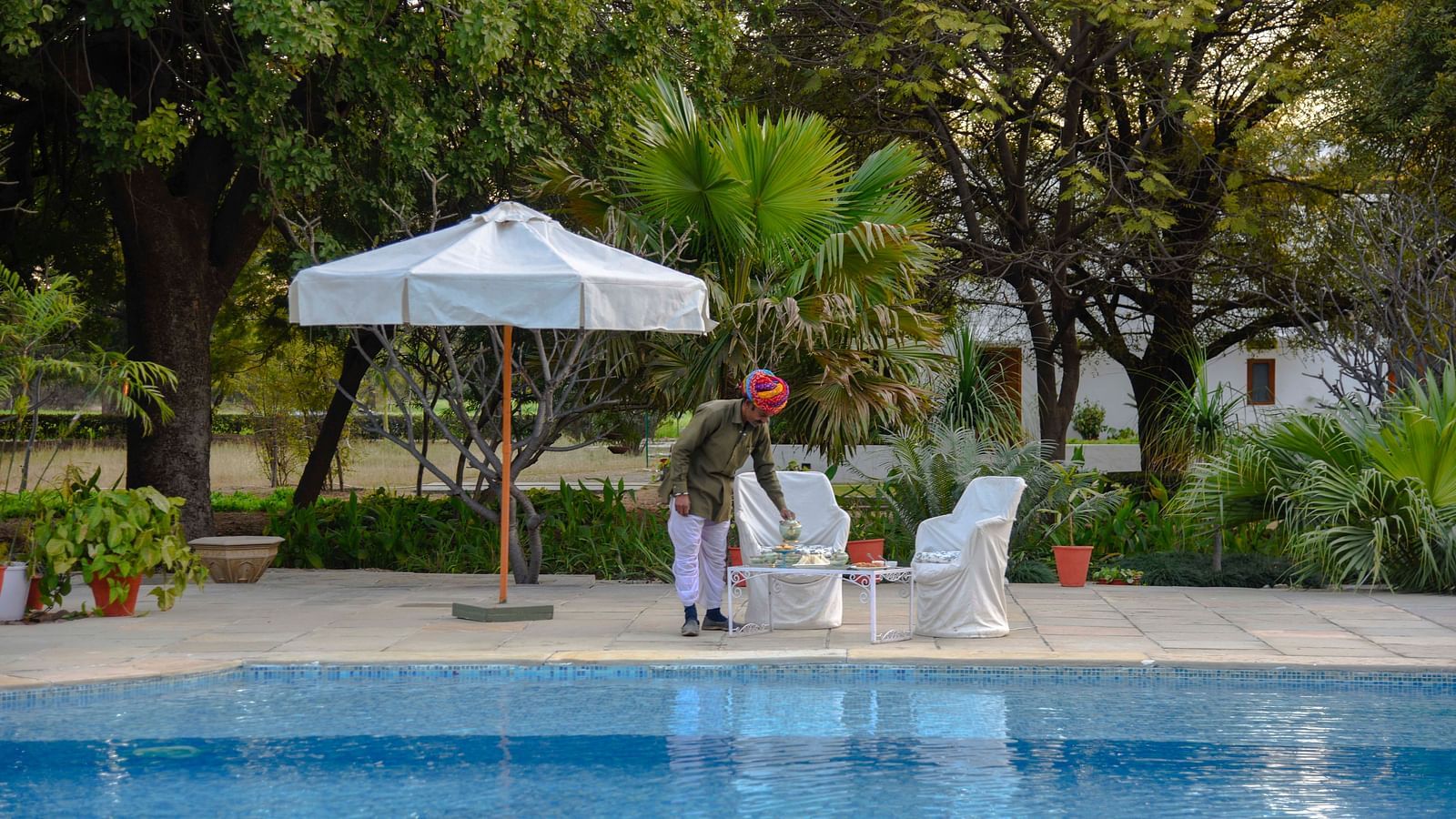 A person in a turban arranges items on a table beside a swimming pool, with a parasol and seating nearby, amidst lush greenery- Shahpura Bagh.