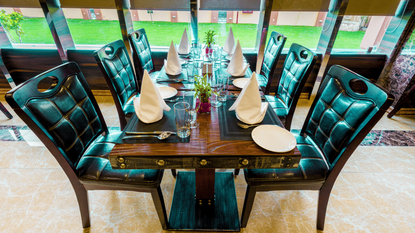 Elegant dining table with four teal tufted chairs and white napkins, set against large windows overlooking lush greenery at Golden Tulip Kukas, Jaipur.