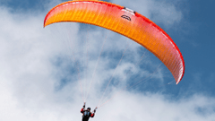 Paraglider flying beneath a bright orange canopy against blue skies, with soft white clouds and rugged mountain terrain far below