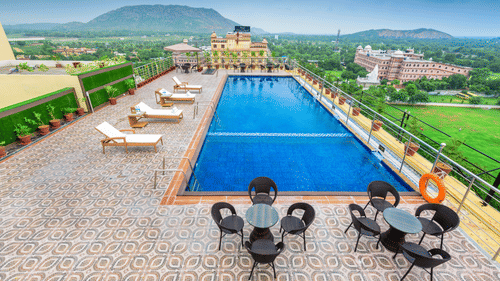 High-angle view of a rooftop pool with sun loungers and patio sets, offering a scenic outlook of lush green plains and distant mountains at Golden Tulip Kukas, Jaipur.