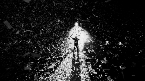 An outdoor concert, with the lead singer under white spotlight and white paper shreds floating in the air.