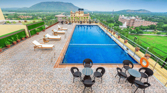 High-angle view of a rooftop pool with sun loungers and patio sets, offering a scenic outlook of lush green plains and distant mountains at Golden Tulip Kukas, Jaipur.