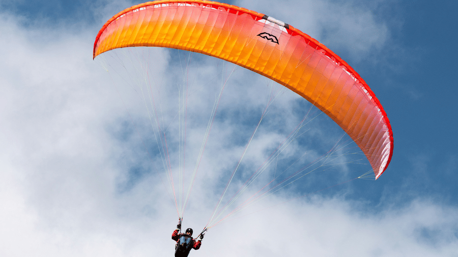 Paraglider flying beneath a bright orange canopy against blue skies, with soft white clouds and rugged mountain terrain far below
