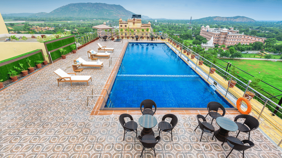 High-angle view of a rooftop pool with sun loungers and patio sets, offering a scenic outlook of lush green plains and distant mountains at Golden Tulip Kukas, Jaipur.