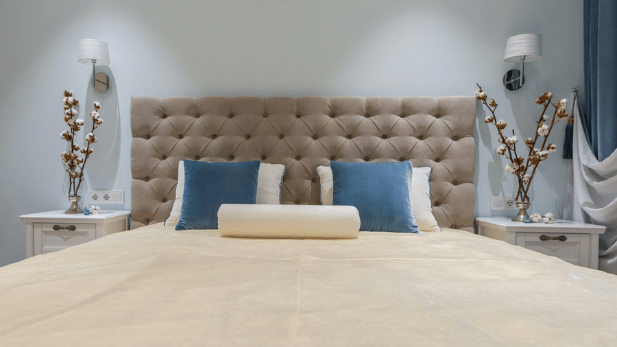 A wide bed features a tan tufted headboard, blue accent pillows and white bedside tables on either side holding decorative floral arrangements in vases.