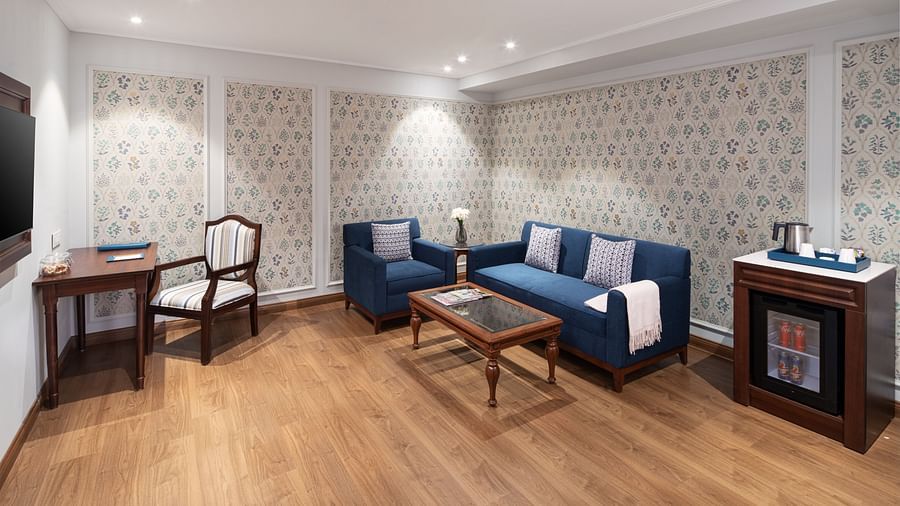 An image of the living room of our Suite in Haridwar with large blue couches, a work desk, a wall-mounted television, and a mini fridge - MJ Sarovar Portico, Haridwar