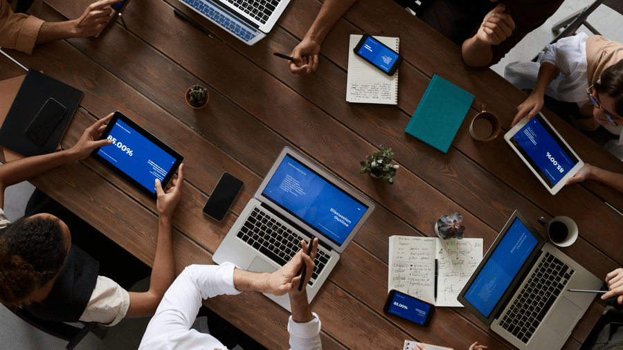 A high-angle view of several people gathered around a conference table working on laptops and phones during a meeting.