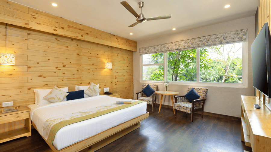 Spacious Villa Grande bedroom with a large bed, wooden walls, a seating area near the window, and a ceiling fan  at The Golden Tusk, Jim Corbett