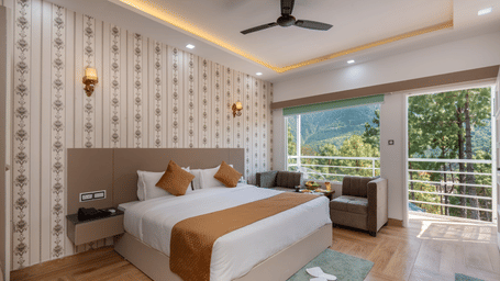 A spacious hotel bedroom featuring a large double bed with crisp white linens and brown accents, a cosy seating area, and a balcony offering scenic views of lush greenery and hills - DLS Nature Trinket Resort, Dalhousie