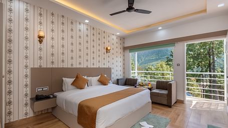 Spacious hotel bedroom featuring a large double bed with crisp white linens and brown accents, stylish wallpaper, a ceiling fan, modern lighting, a cosy seating area, and a balcony offering scenic views of lush greenery and hills - DLS Nature Trinket Resor