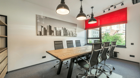 A modern conference room with a large wooden table, several black office chairs, hanging industrial-style pendant lights, and a window with red blinds.