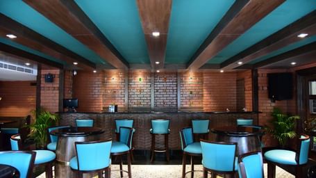 Another view of the bar at The Royal Comfort, highlighting the long wooden bar top, the brick wall behind it, and the stylish blue-green chairs.