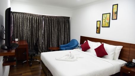  A spacious royal deluxe room at The Royal Comfort, featuring a king-sized bed with rich wooden accents, a deep red runner, and a modern painting above the headboard.