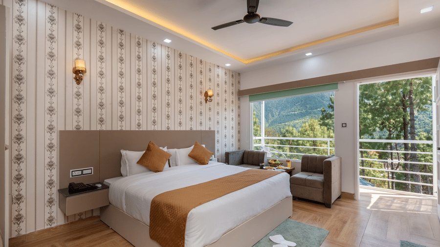 A spacious hotel bedroom featuring a large double bed with crisp white linens and brown accents, a cosy seating area, and a balcony offering scenic views of lush greenery and hills - DLS Nature Trinket Resort, Dalhousie