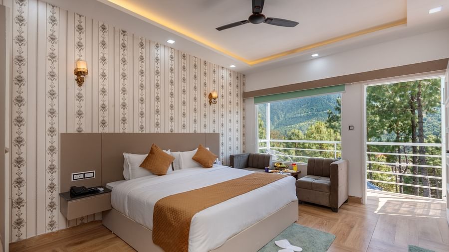 Spacious hotel bedroom featuring a large double bed with crisp white linens and brown accents, stylish wallpaper, a ceiling fan, modern lighting, a cosy seating area, and a balcony offering scenic views of lush greenery and hills - DLS Nature Trinket Resor