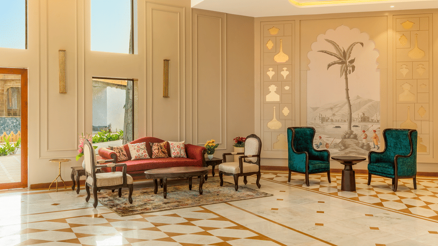 An indoor sitting area at EsthereaRaj Leela, Ranakpur, featuring a red sofa, teal velvet chairs, and light-coloured walls with subtle artwork.