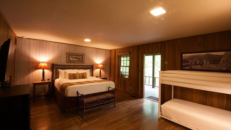 a dimly lit room featuring a double bed along with a bunk bed in the corner with a door opening to the exterior of the cabin - Historic Tapoco Lodge