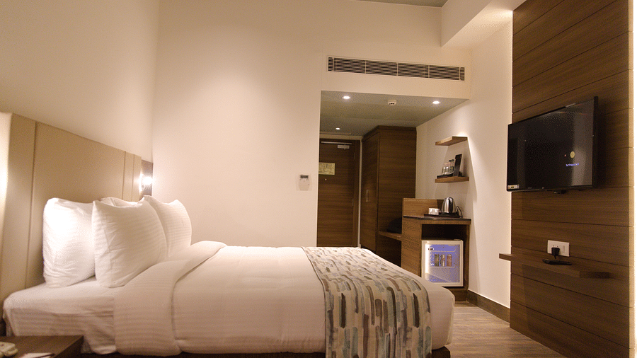 A modern hotel room at Lords Inn Udaipur offers a comfortable bed with white linens, a wall-mounted TV, and subtle lighting.