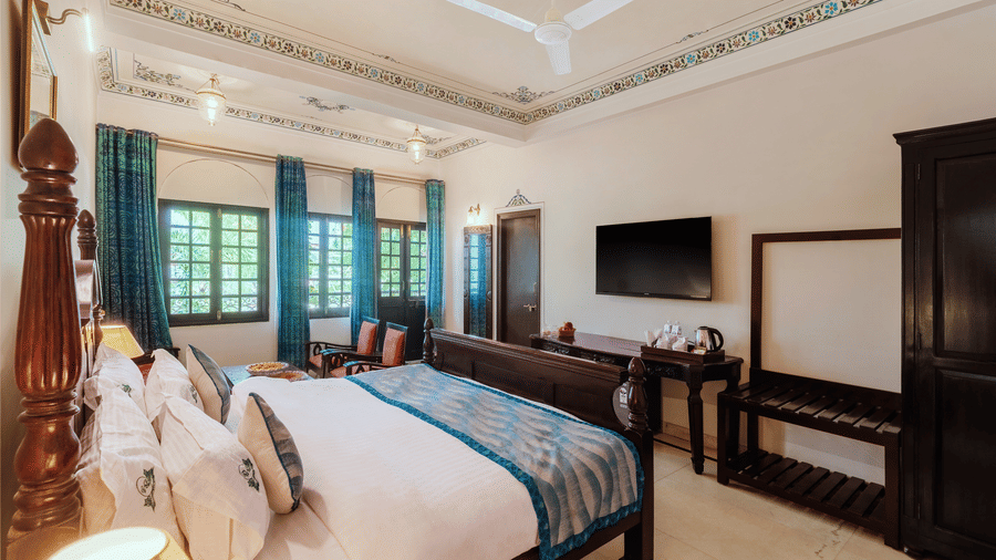 A view from the headboard in a spacious bedroom at Panna Vilas Palace, showcasing a wall-mounted television above a wooden table, with a window beside it and curtains pulled to the side | Panna Vilas Udaipur