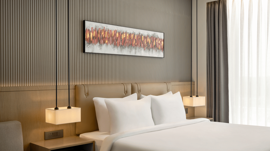 Close-up view of a king size bed with crisp white linens, warm pendant lights, a wooden bedside table, and a decorative wall painting above.