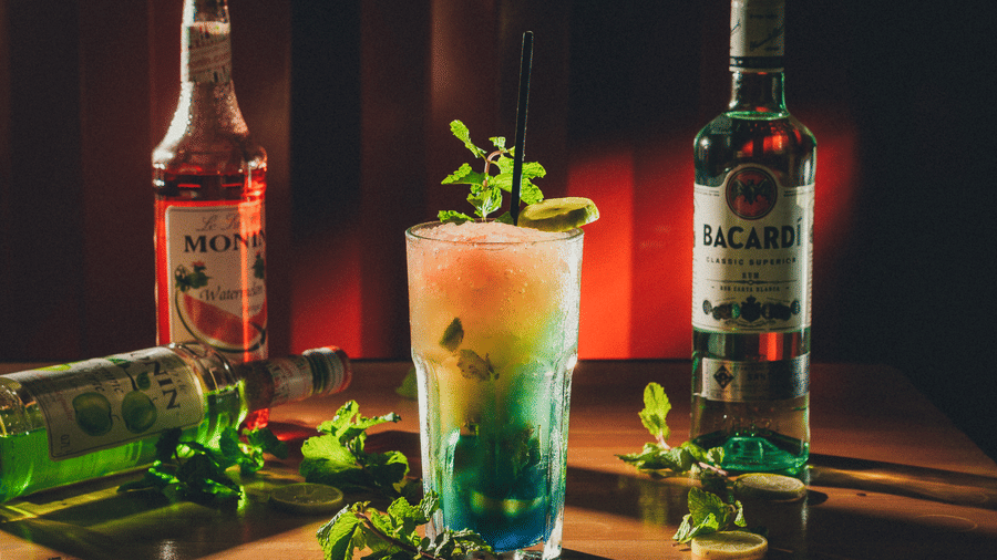 A blue and green layered cocktail is visible next to several bottles of alcoholic spirits and fresh herbs on a wooden surface at S Hotels, Chennai.