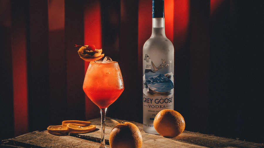 An orange coloured cocktail sits next to a bottle of spirit and citrus fruits on a wooden surface against a dark red backdrop at S Hotels, Chennai.