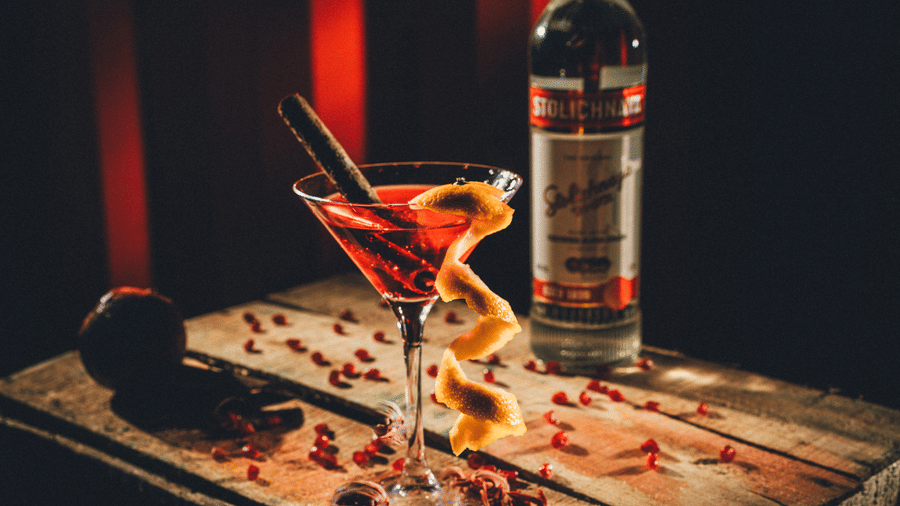 A red martini cocktail in a glass sits next to a bottle of spirit on a wooden surface garnished with coffee beans at S Hotels, Chennai.