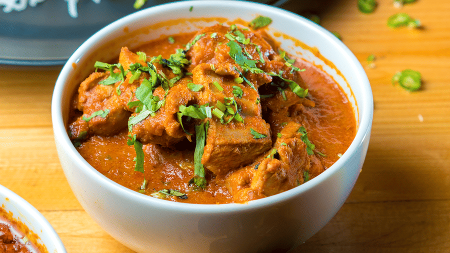 A close up of a bowl of meat curry with herbs on top is visible next to a dark plate at S Hotels, Chennai.