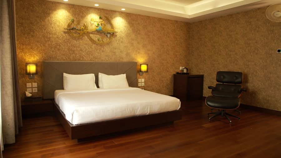 A warmly lit bedroom with a large bed, decorative wall panel, bedside lamps, and wooden flooring at The Hamuse Hotel, Kodaikanal.