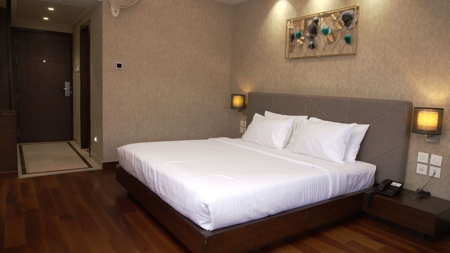A spacious bedroom with a large bed, wall art above the headboard, bedside lamps, and wooden flooring at The Hamuse Hotel, Kodaikanal.