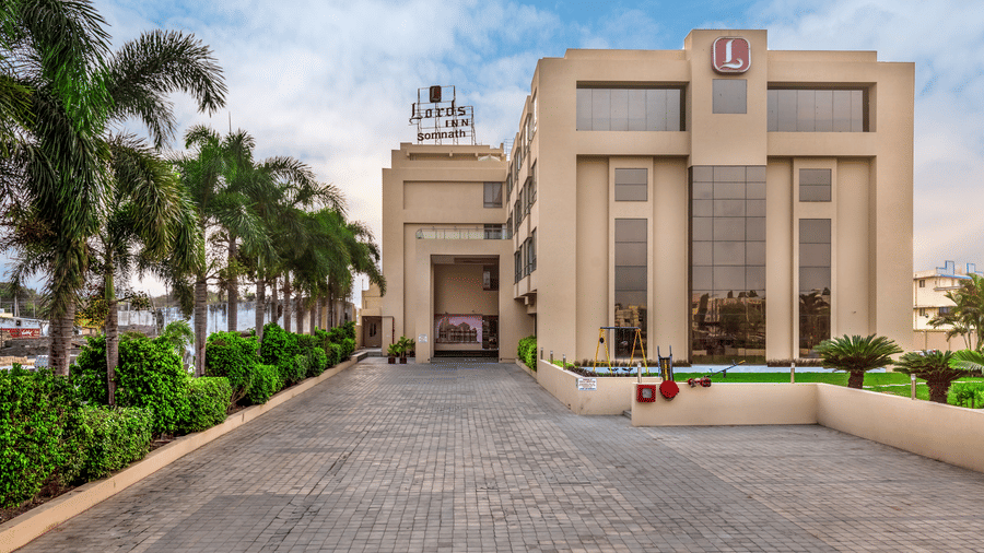 A wide shot of the facade of Lords Inn Somnath with a prominent entrance and surrounding greenery, including paved walkways and some parked cars, under a partly cloudy sky.