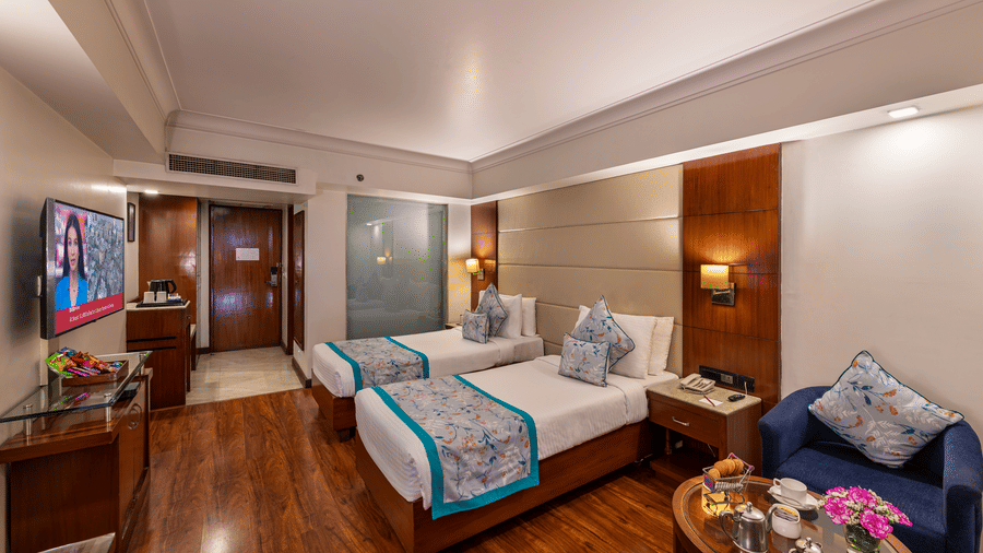 Wide view of deluxe twin bedroom from a different angle with wooden flooring, flat screen TV, sitting area and bright lighting Lords Plaza Jaipur.