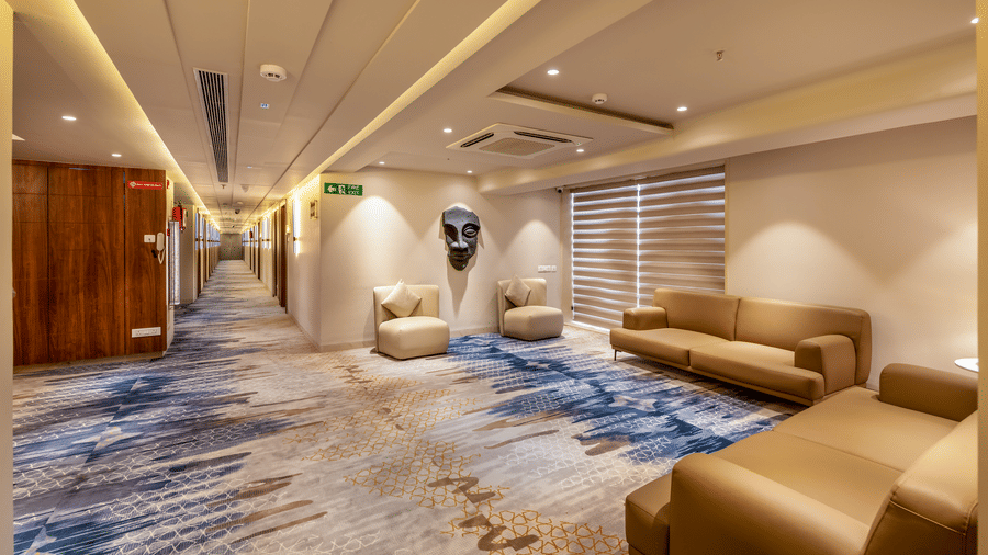 A brightly lit hotel corridor and lounge area with tan leather sofas, a patterned blue and gold carpet, and a decorative wall mask - Skkylight Lords Inn, Nanded