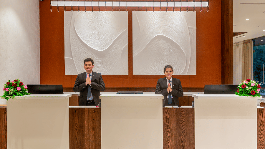 2 receptionists in suits greeting with their hands joined together behind a wooden front desk with white panels, featuring an orange wall and textured art - Skkylight Lords Inn, Nanded