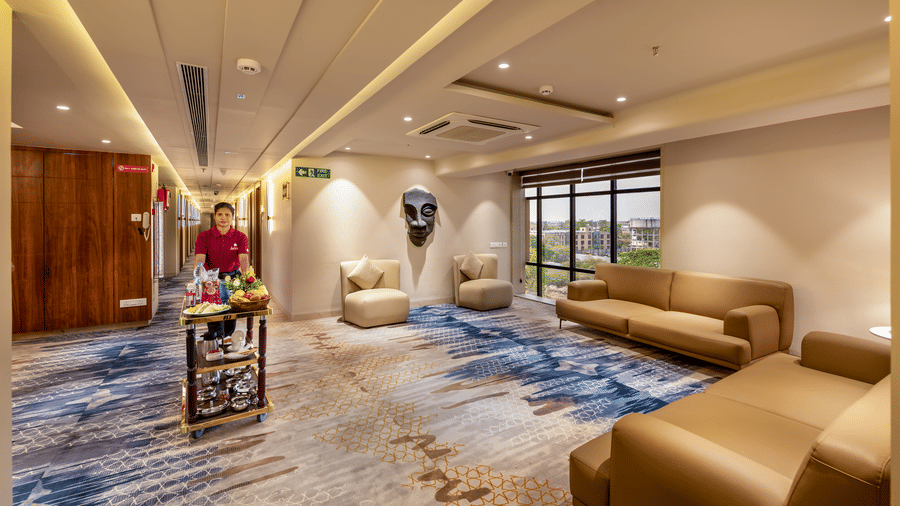  A hotel staff member pushes a wooden food trolley laden with fruit and refreshments through a modern lounge area with blue patterned carpet - Skkylight Lords Inn, Nanded