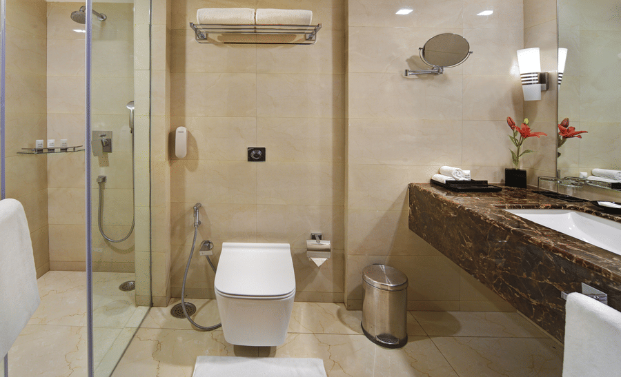 Bathroom at The Bristol Hotel, Gurgaon with beige marble tiles, a glass-enclosed shower, a wall-mounted toilet, and a dark marble vanity counter with a mirror.