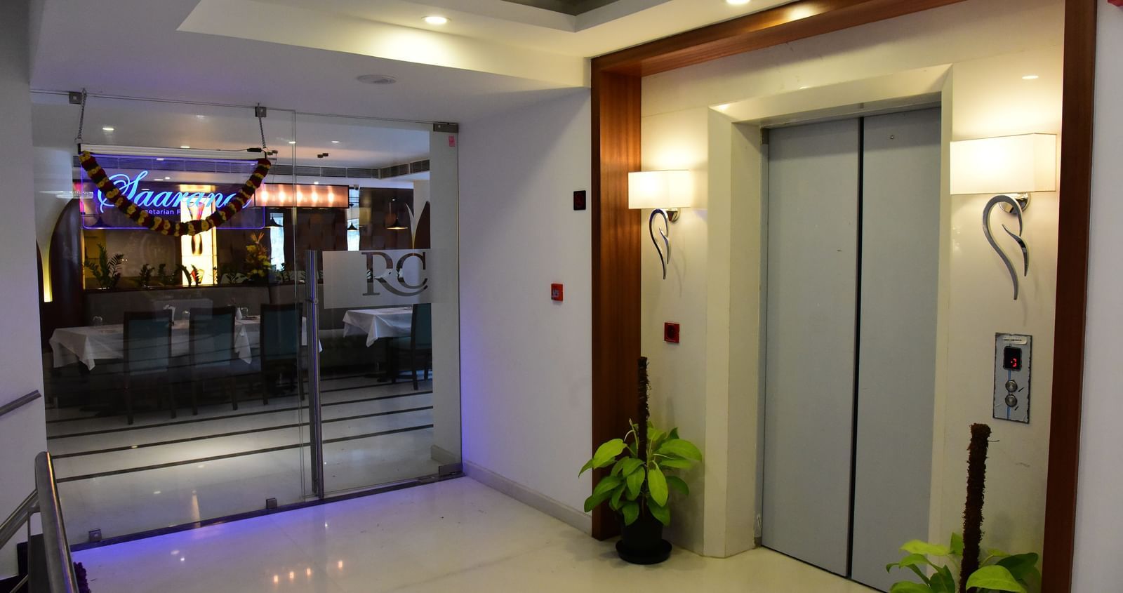 A sleek, modern elevator lobby at The Royal Comfort, with a metallic elevator door and glowing blue lights beneath a wooden ceiling panel.