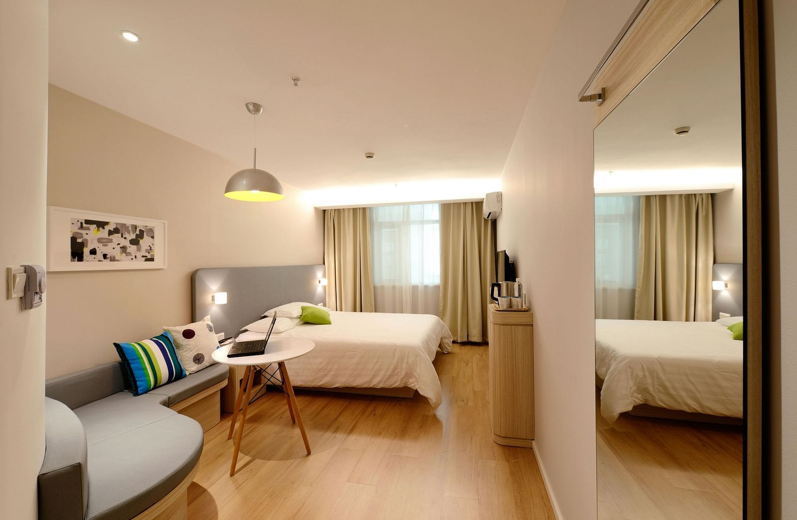 A bright hotel suite includes a seating area and a bed, reflected in a large mirror.