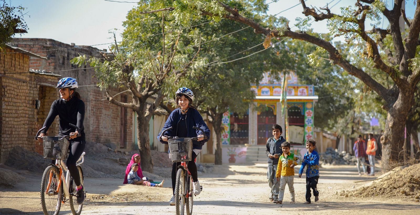 An image of two children cycling with buildings and people behind them- Shahpura Bagh.