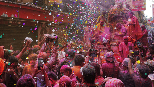 A crowd celebrating Holi on a narrow street, with people throwing coloured powder and flower petals into the air.
