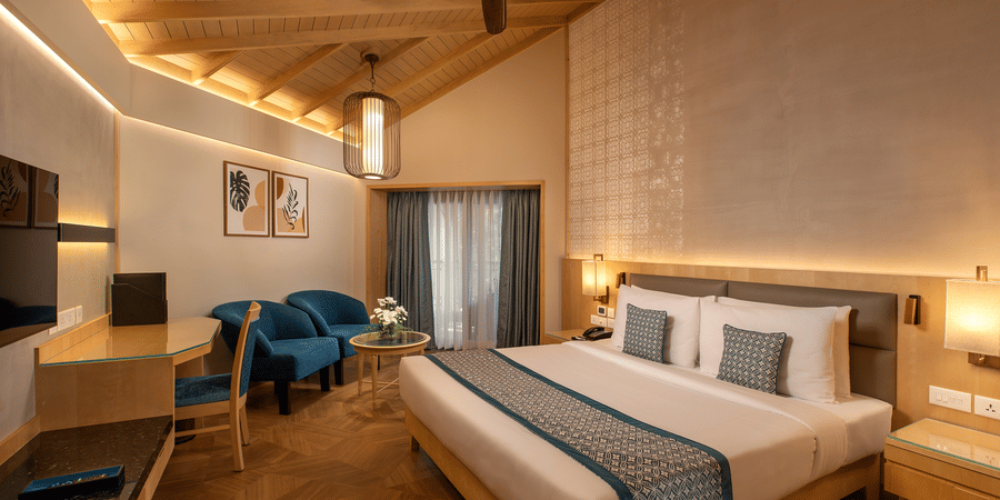 A view of the Superior Deluxe Room with a king bed, side tables, lamps, a tv, ceiling fan, chairs in the corner with a coffee table at Kenilworth Resort & Spa, Goa, one of the best workation places in Goa.