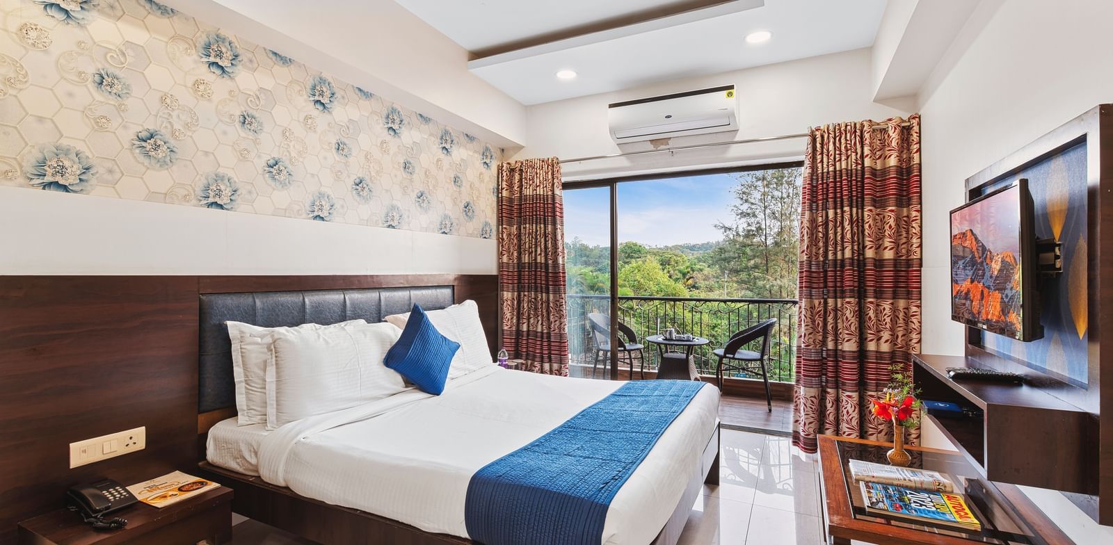Bright and ventilated room at Mayur Retreat with a blue-themed bed, floral walls, and balcony garden view.