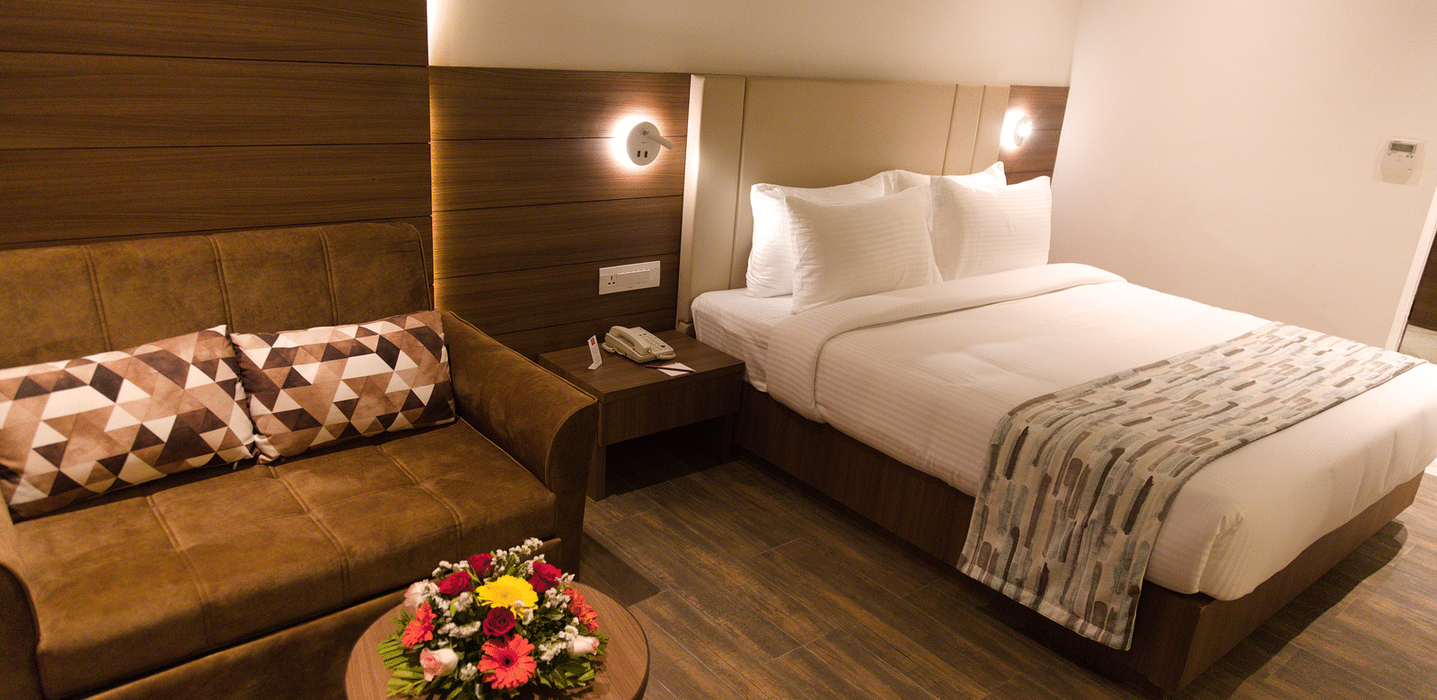 A well-appointed hotel room at Lords Inn Udaipur features a comfortable bed, a cozy sofa with patterned pillows, and a small round table with flowers.