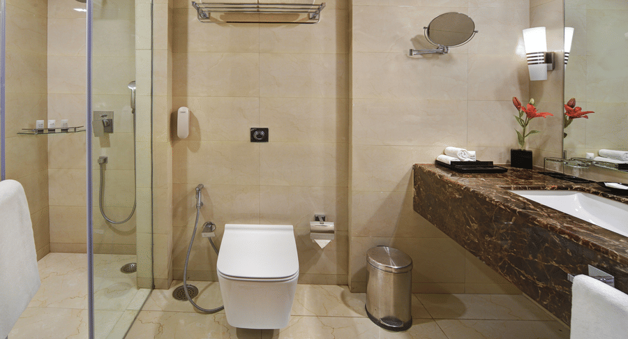 Bathroom at The Bristol Hotel, Gurgaon with beige marble tiles, a glass-enclosed shower, a wall-mounted toilet, and a dark marble vanity counter with a mirror.