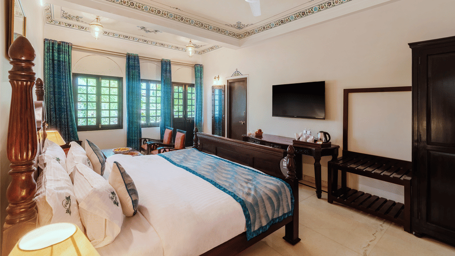 A view from the headboard in a spacious bedroom at Panna Vilas Palace, showcasing a wall-mounted television above a wooden table, with a window beside it and curtains pulled to the side | Panna Vilas Udaipur