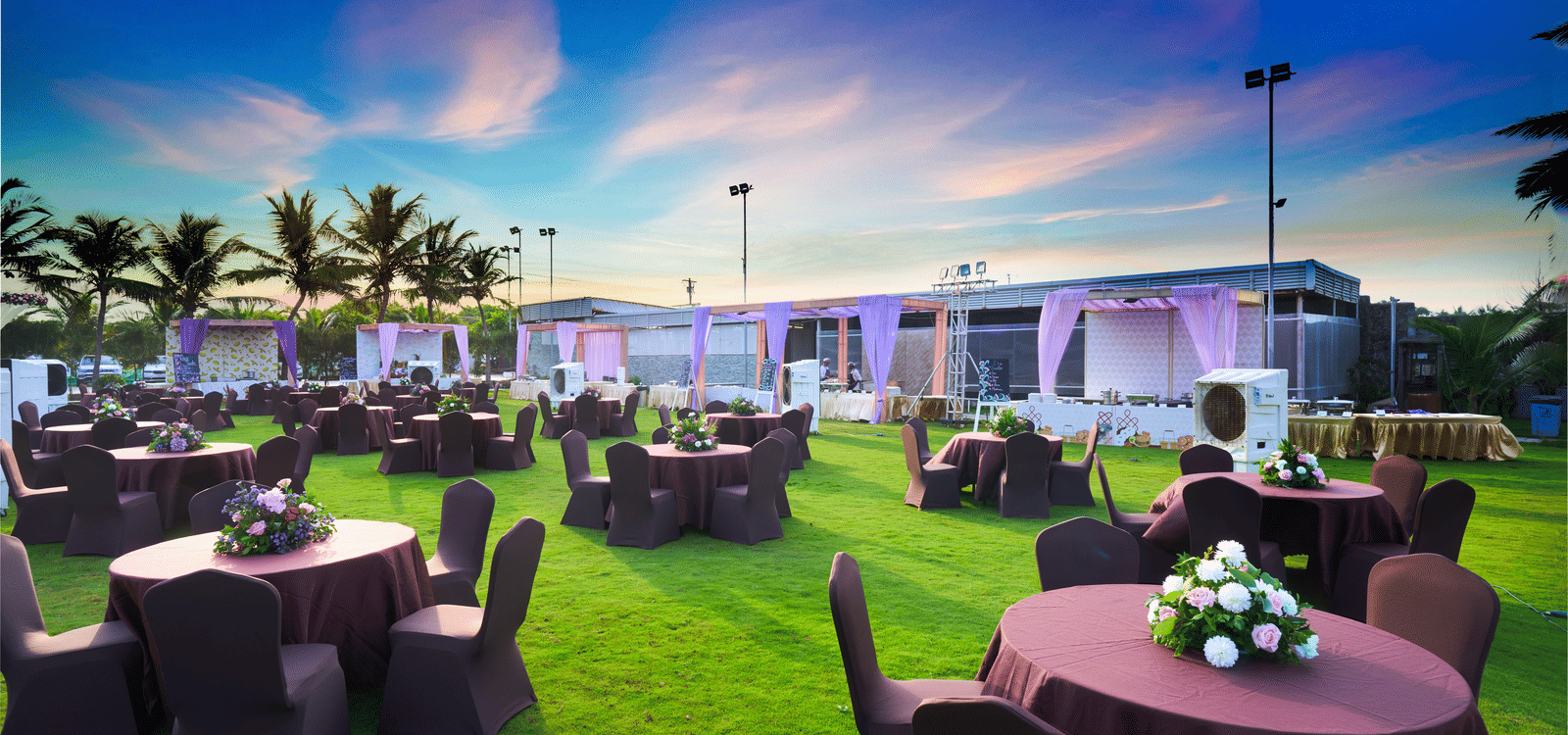 Outdoor lawn setup with round tables and floral centrepieces under a vibrant evening sky - Grande Bay Resort & Spa, Mamallapuram