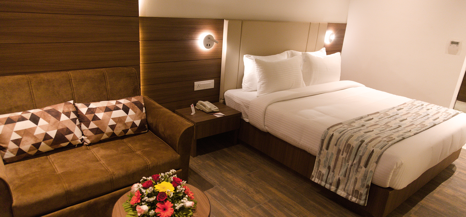A well-appointed hotel room at Lords Inn Udaipur features a comfortable bed, a cozy sofa with patterned pillows, and a small round table with flowers.