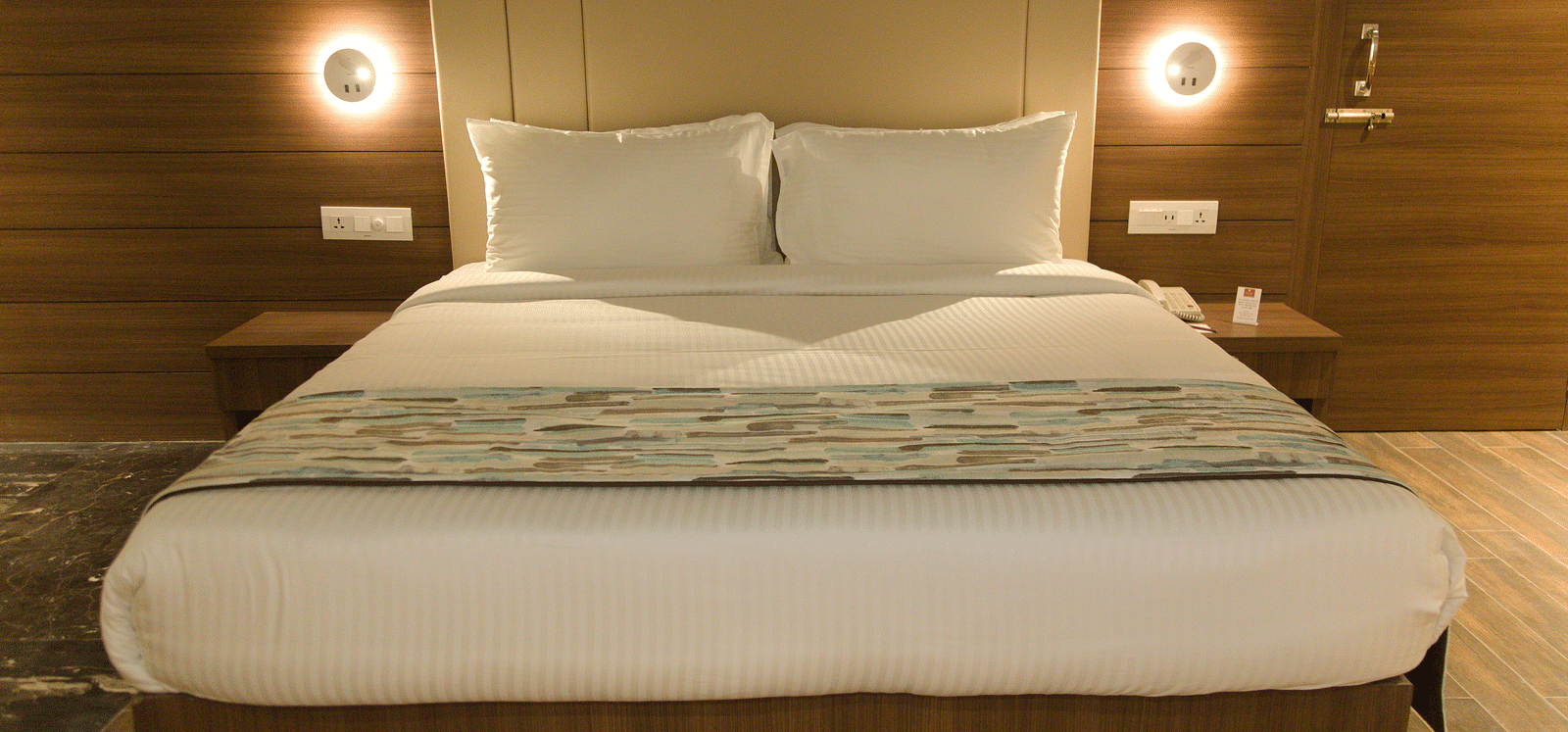 A neatly made bed with white linens and a patterned runner is centered between two illuminated wall sconces and bedside tables.